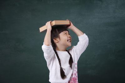 Cheerful girl holding book on head while standing by blackboard