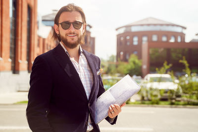 Portrait of smiling businessman holding document while standing outdoors