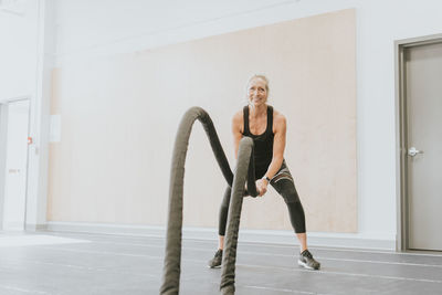 Full length portrait of smiling mature woman exercising with ropes at gym