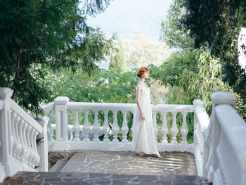 Woman standing on white railing against trees