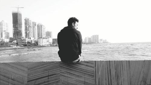 Rear view of man sitting on retaining wall by sea against sky