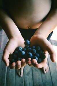 Low section of shirtless boy holding blueberries while standing on hardwood floor