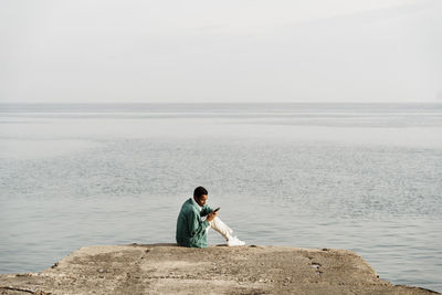 Young man using mobile phone while sitting on pier by sea and sky