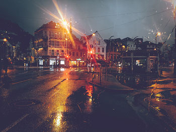 Illuminated street amidst buildings against sky during rainy season at night