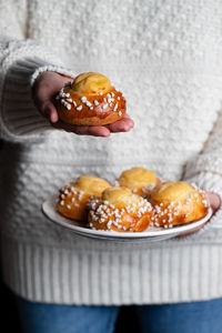 Crop female in white sweater and jeans holding plate with homemade pastry and demonstrating tasty fresh buns with white sprinkles