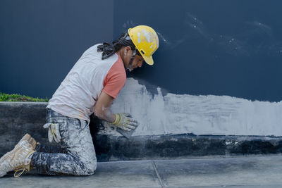 Side view of professional male painter in yellow hardhat in process of plastering wall while sitting on sidewalk on city street