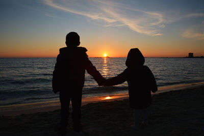 Silhouette siblings holding hands while standing at beach during sunset