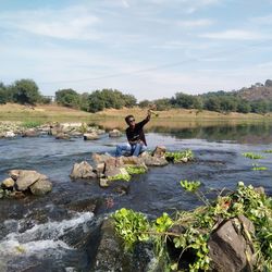 Man throwing plant while sitting on rock in river against sky