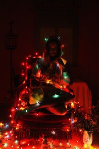 Full length of woman with illuminated string lights sitting on chair at home