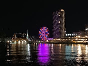 Illuminated ferris wheel by river against sky at night