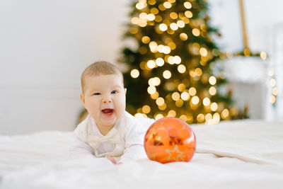 The baby laughs, covering one eye, lying on the bed against the background of christmas lights