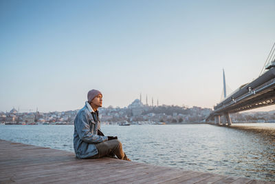 Portrait of young man sitting on pier against clear sky