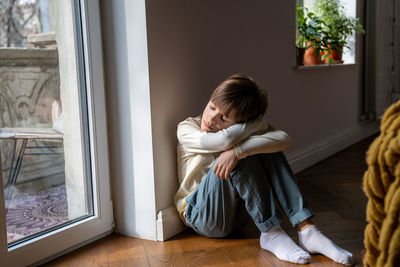 Tired sleepy teen boy sitting on floor embracing knees at home near window.