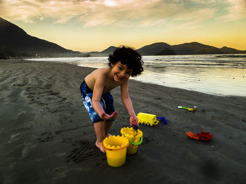 Portrait of smiling boy playing with sand and toys at beach against cloudy sky during sunset