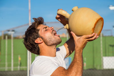 Young man drinking water from pot at court during sunny day