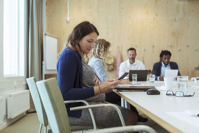 Pregnant businesswoman using mobile phone while sitting with colleagues in board room