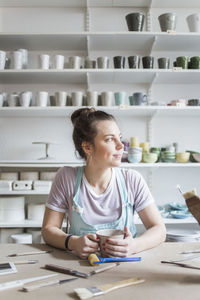 Young female potter sitting at workbench while looking away against shelves at store