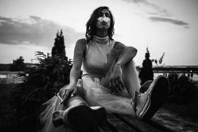 Young woman smoking while sitting on bench against sky