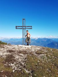 Shirtless man standing by cross on mountain against clear blue sky