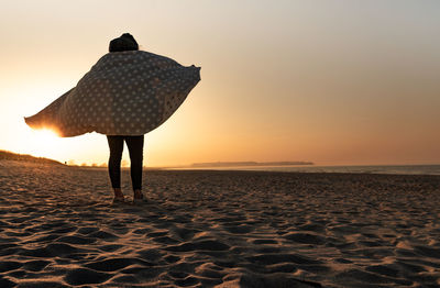 Rear view of woman standing at beach against sky during sunset