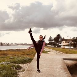 Full length portrait of girl kicking on footpath against cloudy sky