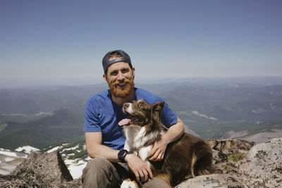 Portrait of man with dog sitting on mountain against sky