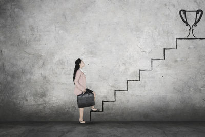 Digital composite image of businesswoman climbing on staircase towards trophy drawn on wall