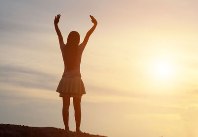 Rear view of woman with arms raised standing against sky during sunset