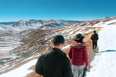 Rear view of three friends walking on snow covered mountain along a dirt path with clear skies