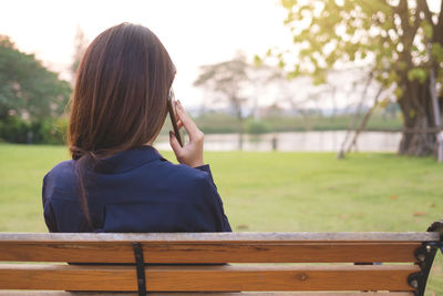 Rear view of woman talking on mobile phone while sitting on bench against trees