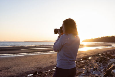 Man photographing at beach against sky during sunset