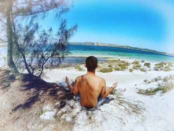 Rear view of shirtless man doing yoga at beach during sunny day