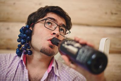 Low angle view of man drinking wine from bottle with grape bunch on ear against wall