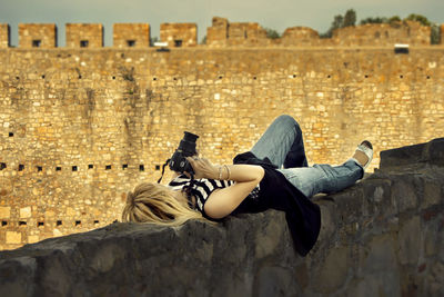 Mature woman with camera lying on retaining wall against old ruin