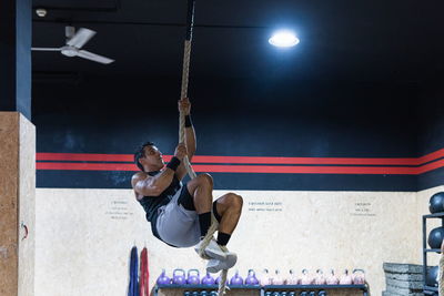 Full body of muscular sportsman in activewear performing exercise on climbing rope during workout in light gym with various equipment