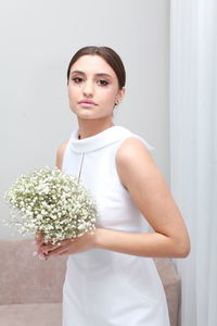 Side view of pretty woman with uptied hairstyle in wedding dress holding minimalist bouquet at home