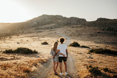 Back view of unrecognizable calm trendy man and woman embracing each other while standing on trail against hillside during sunset