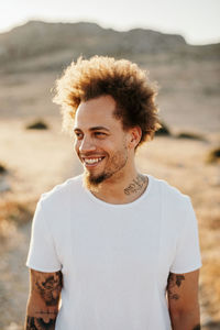 Cheerful trendy male with tattoo in casual clothes standing on dirt road and smiling looking away against blurred rocky hill in sunny evening