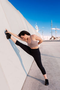 Full body of sporty female in activewear with eyes closed and raised arm doing split and stretching leg during outdoor training against blue sky on summer day