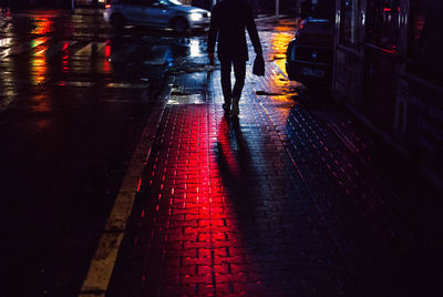 Low section of silhouette man walking on wet sidewalk at night