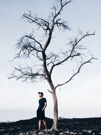 Young man with hands in pockets standing by bare tree on field against sky