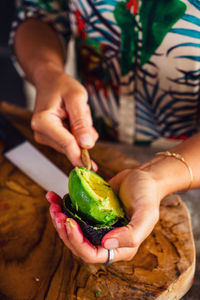 From above anonymous woman using spoon to remove peel from half of ripe avocado while preparing vegan food for lunch