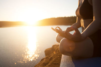 Midsection of woman meditating while sitting on rock at lakeshore during sunset