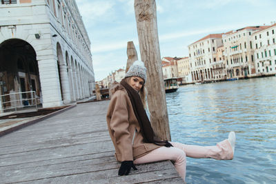 Portrait of young woman sitting on pier over river against historic buildings
