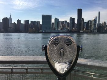 Coin-operated binoculars by river and city skyline against sky