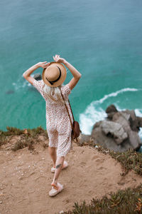 Rear view of woman wearing hat standing on cliff by sea