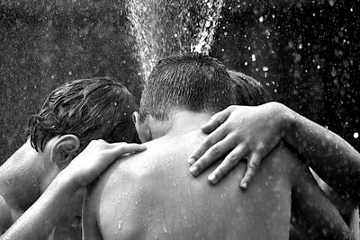 Shirtless friends huddling while enjoying sprinkler in backyard