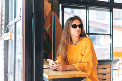 Young woman with long blonde hair wearing sunglasses and yellow longsleeve with mobile phone in cafe