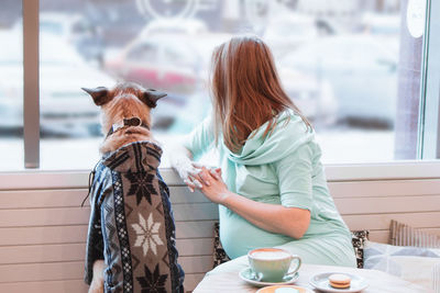 Young pregnant women is sitting in cafe with her dog in sweater and looking into the window.