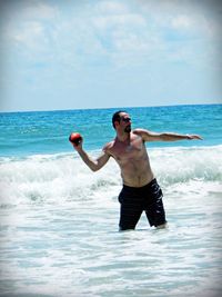 Shirtless man throwing ball while standing in sea against sky
